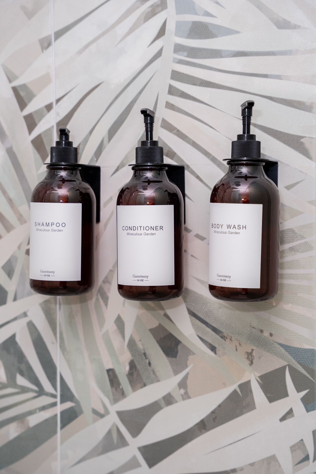 Three bottles of hair care products labeled 'SHAMPOO,' 'CONDITIONER,' and 'BODY WASH' from 'Miraculous Garden' by 'Gaontssny' on a patterned wall.