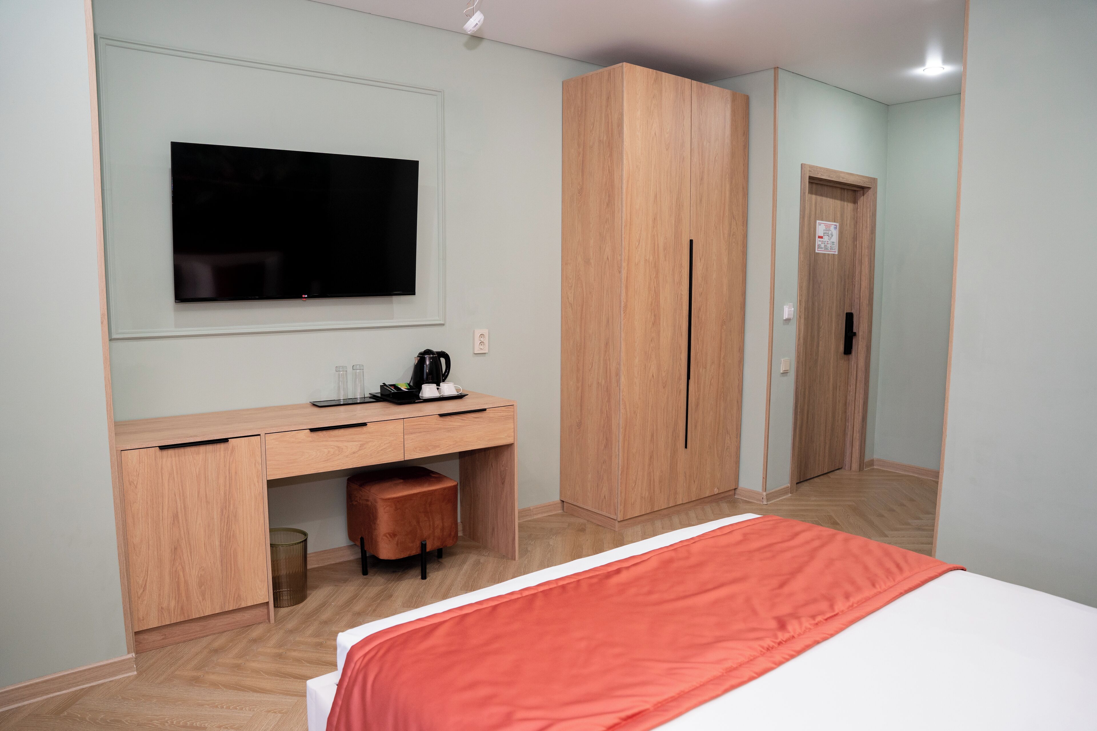 A hotel room with a bed, a desk with a flat-screen TV, a wardrobe, and a door.