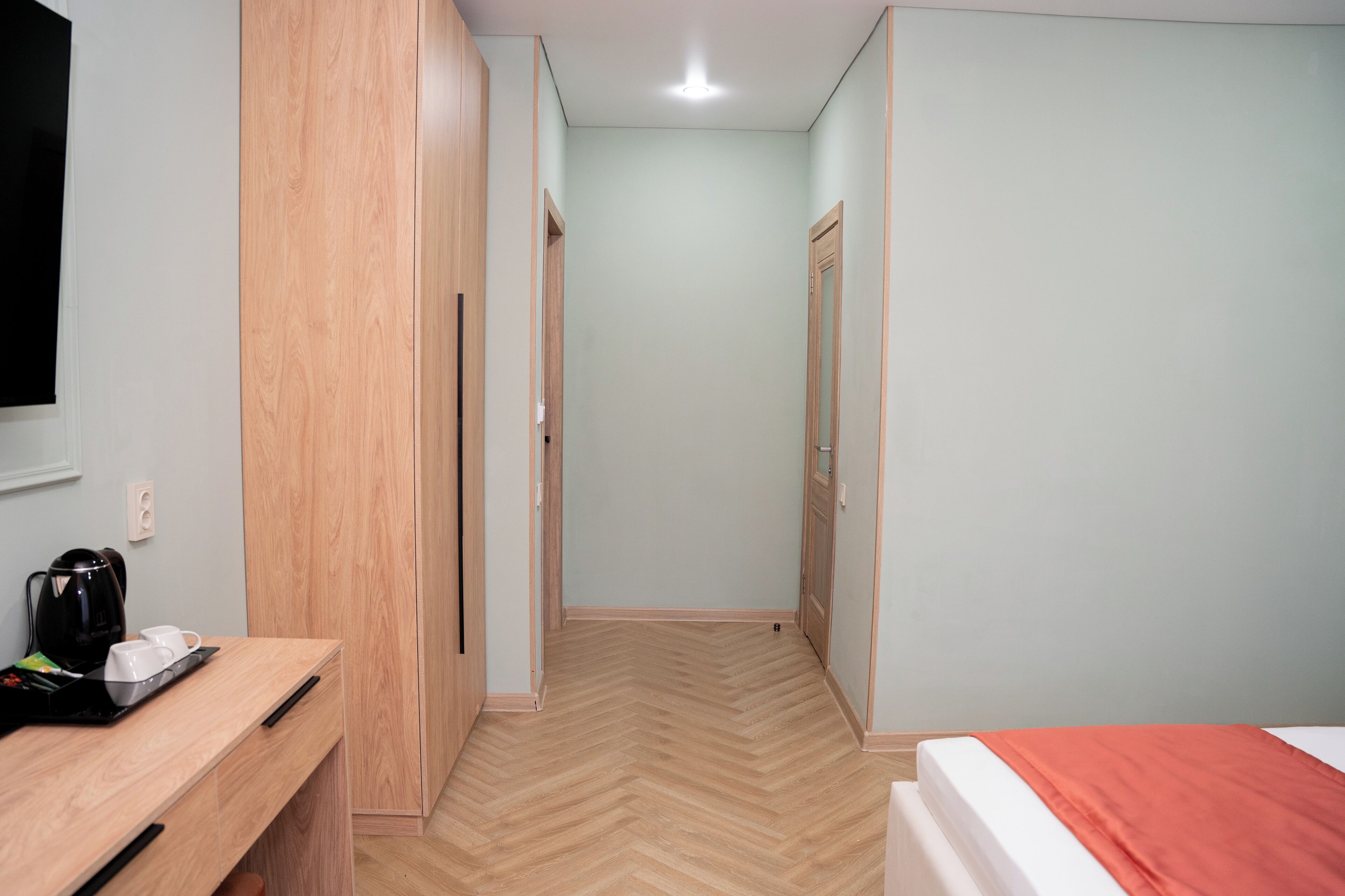 A hallway with a wooden wardrobe, a bed with a red blanket, and a coffee maker on a desk.