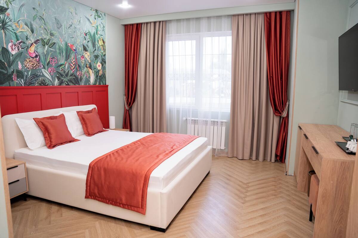 A hotel room with a large bed, a wooden desk, a television, and a window with curtains.