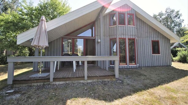 Une maison moderne avec un toit en pente, de grandes fenêtres et une terrasse en bois équipée d’une table et de chaises.