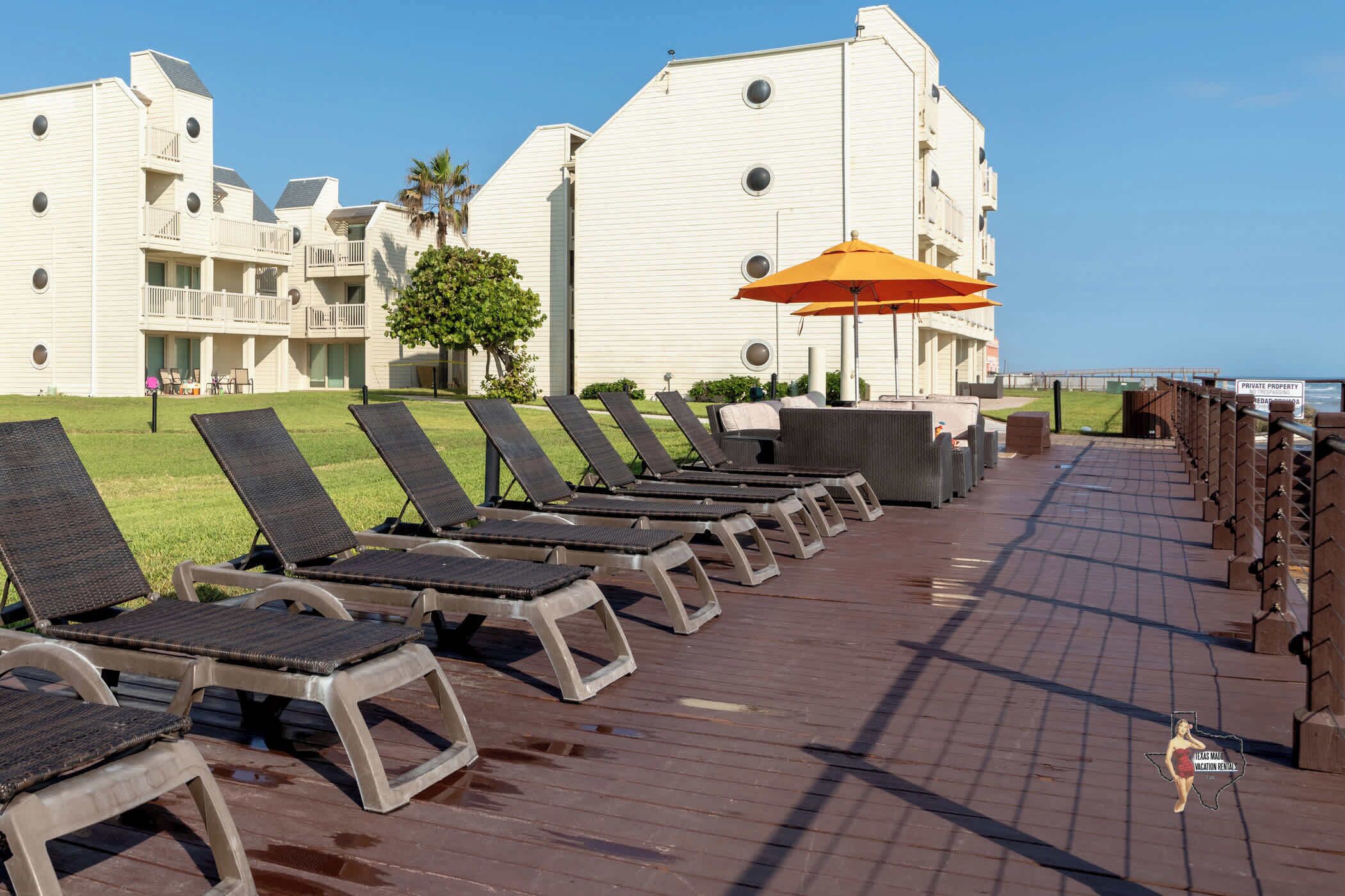 A row of sun loungers on a wooden deck with a view of a white building and the ocean.