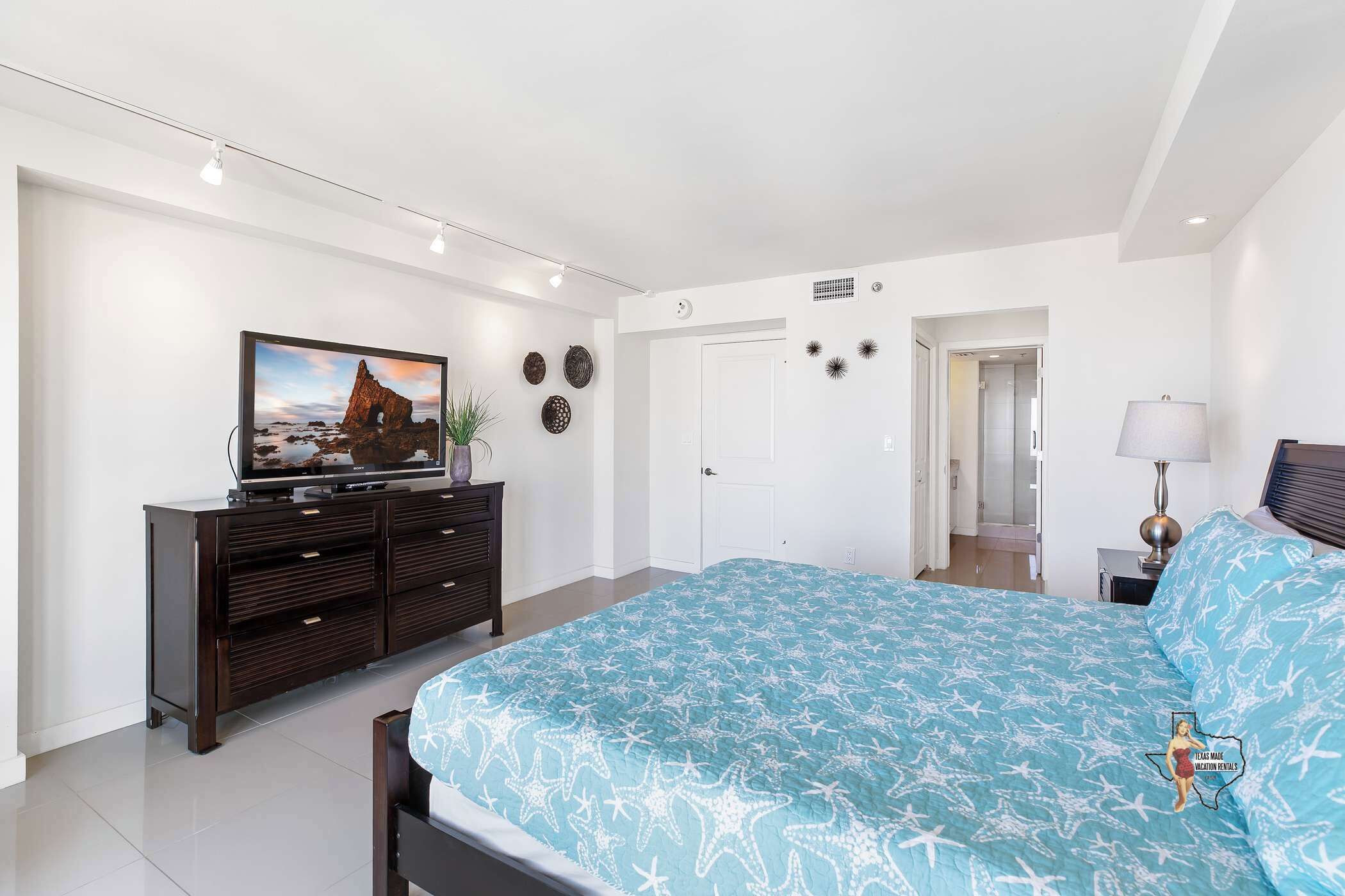Een moderne slaapkamer met een groot bed, een televisie op een donkere dressoir en een deur naar een andere kamer.