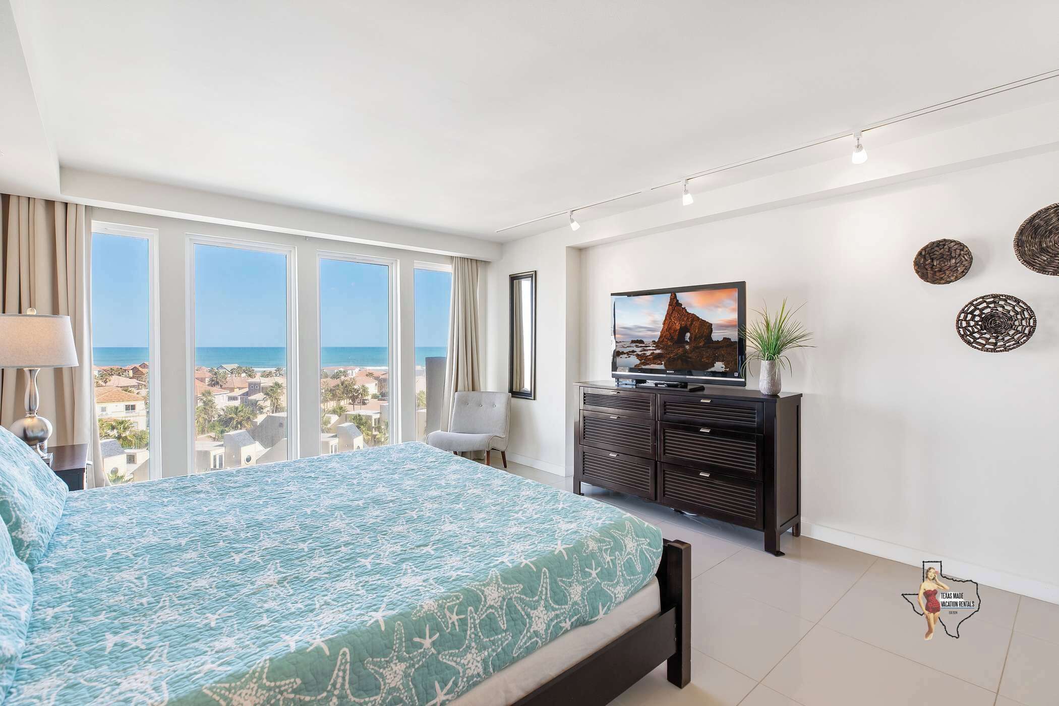 A bedroom with a large bed, a TV, a dresser, and a view of the sea.