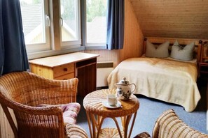 Ein gemütliches Schlafzimmer mit einem Bett, einem Korbstuhl, einem kleinen runden Tisch mit Teekanne und Tasse sowie einem Fenster mit Vorhängen.
