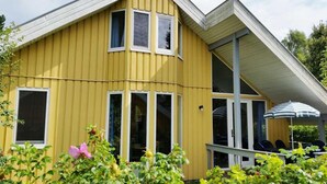 Keltainen rakennus, jossa on kalteva katto, suuret ikkunat ja katettu ulkotila, jossa on istuimia ja aurinkovarjo.