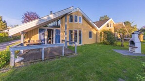 Une maison en bois à deux étages, avec un toit en pente, une véranda dotée de sièges extérieurs et un barbecue dans le jardin.