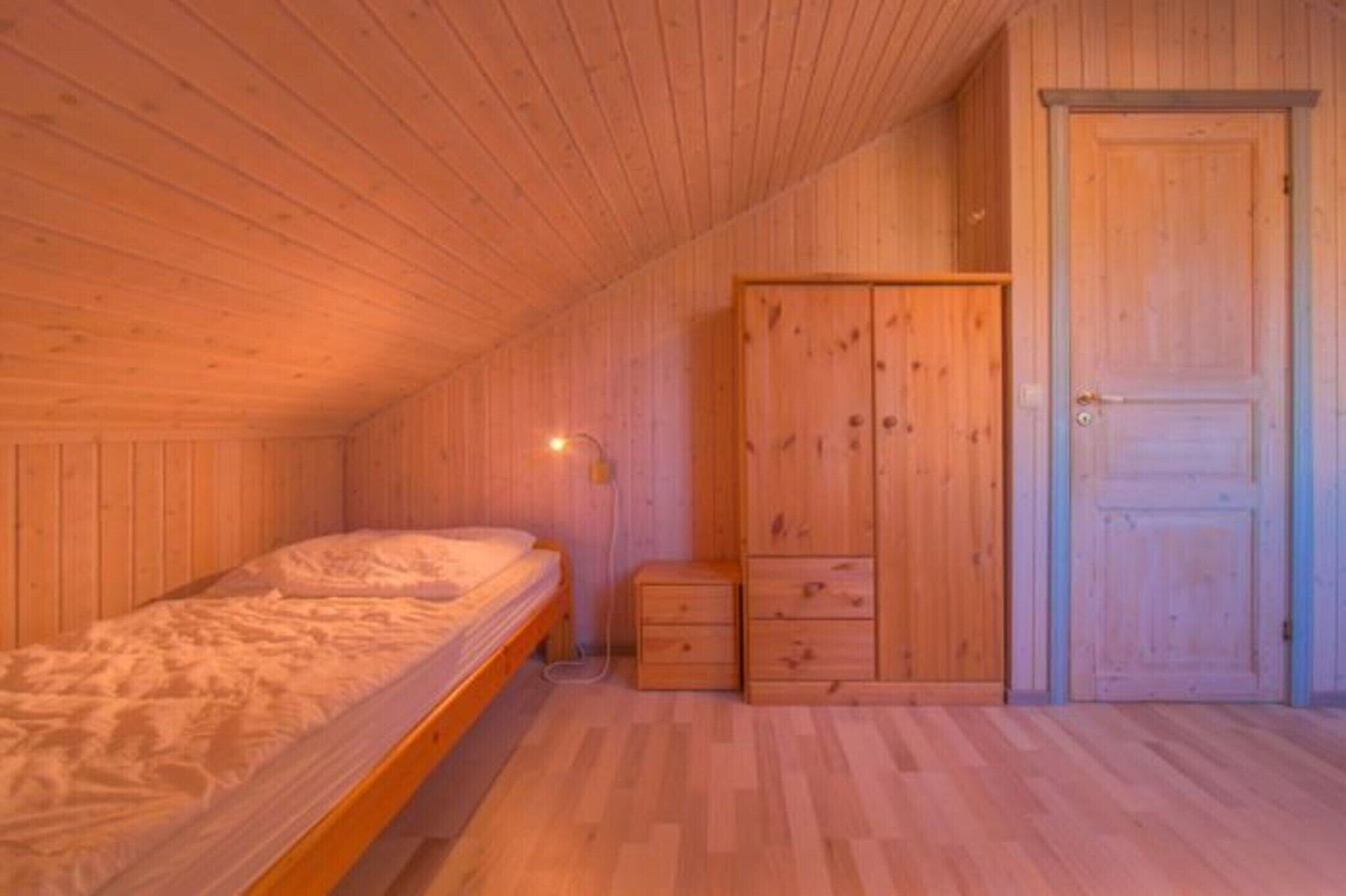 A room with wooden walls and ceiling, a single bed with a white mattress, a wooden wardrobe, and a door.
