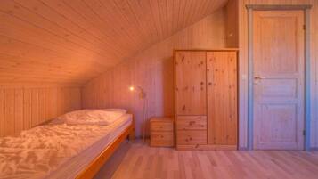 A room with wooden walls and ceiling, a single bed with a white mattress, a wooden wardrobe, and a door.