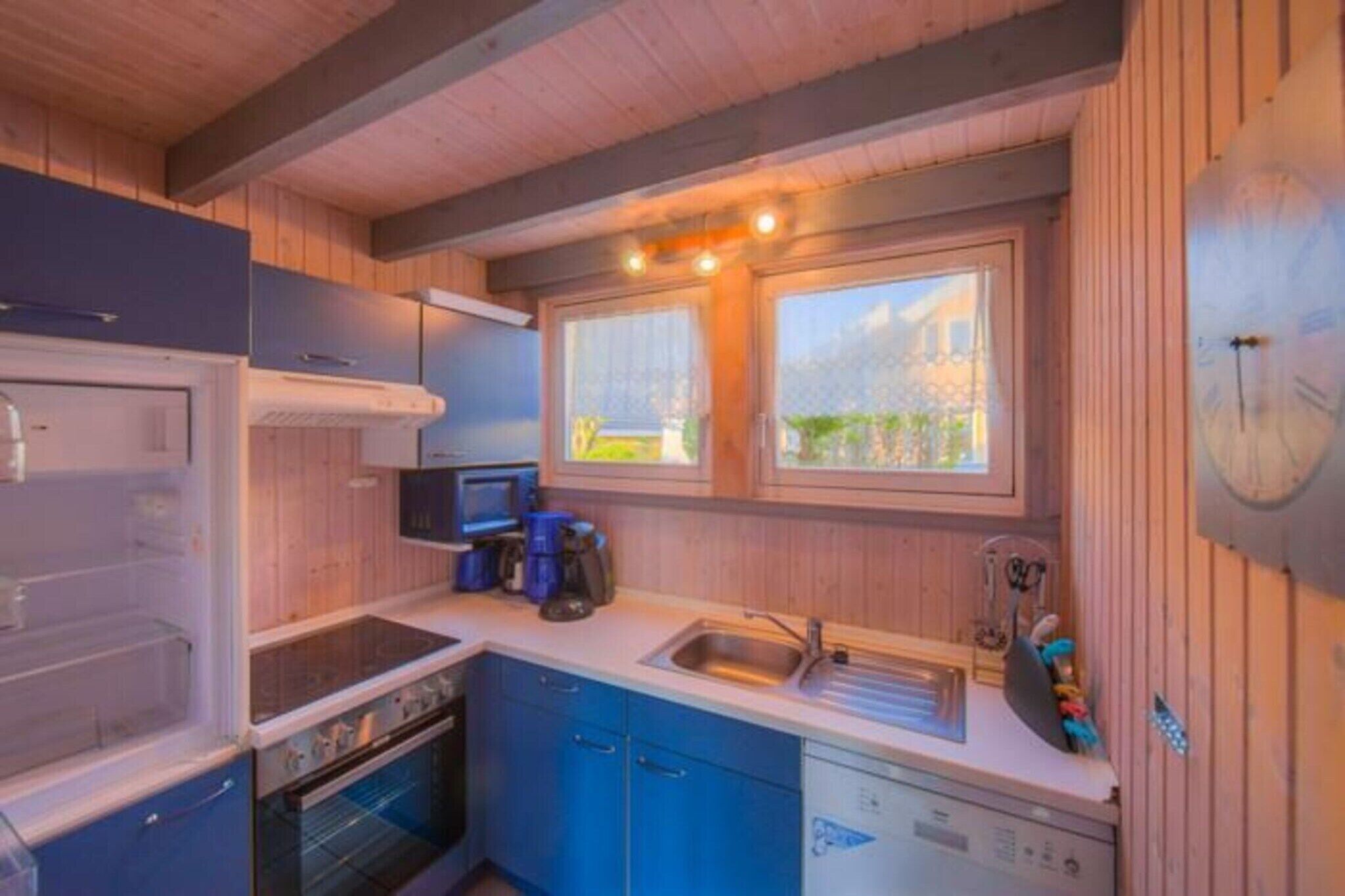 A compact kitchen with a stove, oven, sink, and blue cabinets. There are two windows providing natural light and a view of greenery outside.