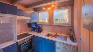 Une cuisine compacte équipée d’une cuisinière, d’un four, d’un évier et de meubles de rangement bleus. Deux fenêtres laissent entrer la lumière naturelle et offrent une vue sur la verdure environnante.
