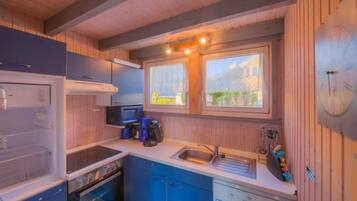 A compact kitchen with a stove, oven, sink, and blue cabinets. There are two windows providing natural light and a view of greenery outside.