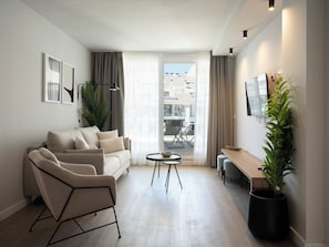 Ein modernes Wohnzimmer mit einer Couch, einem Sessel, einem kleinen runden Tisch, einer Topfpflanze und einem an der Wand befestigten Fernseher.