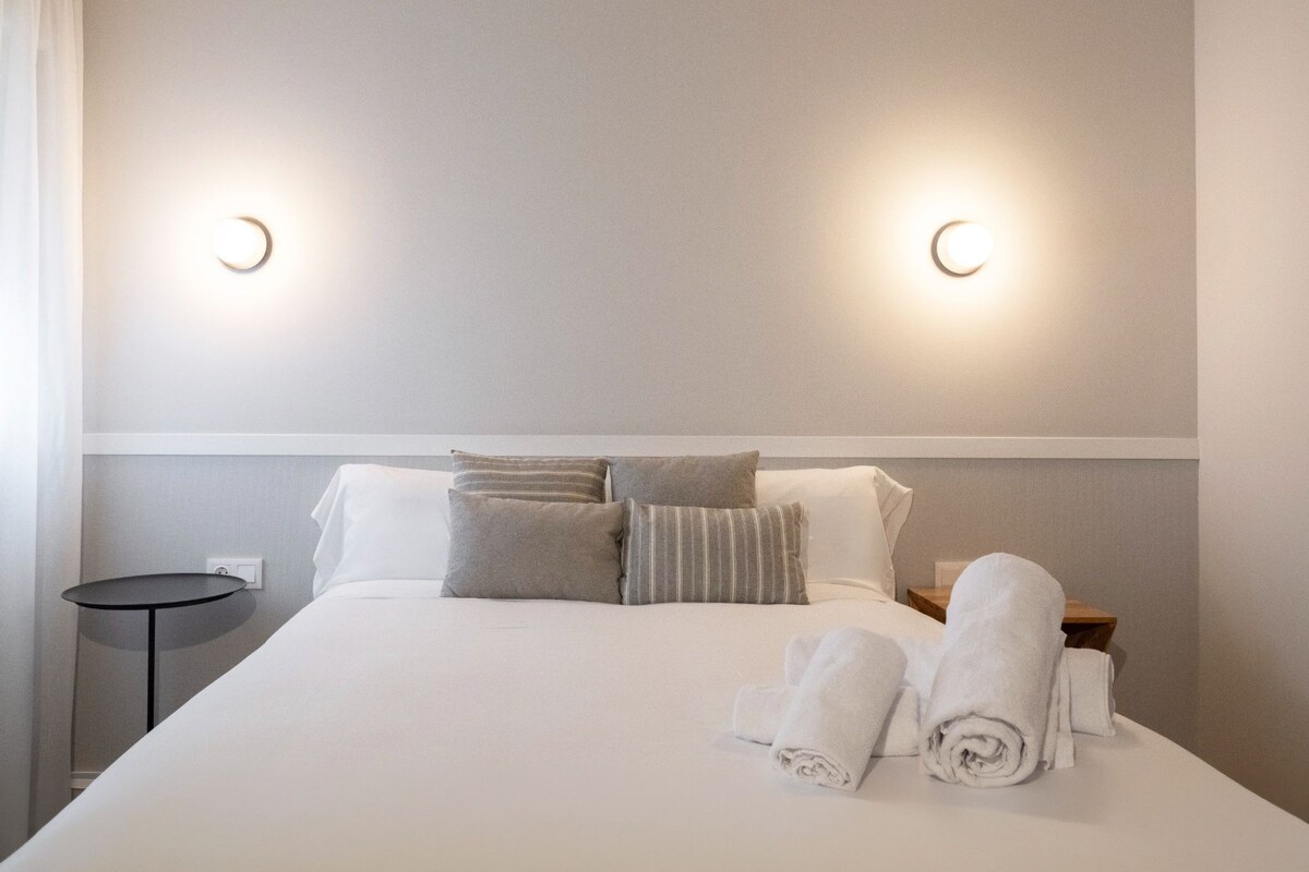 Una habitación de hotel con cama, almohadas, mesita de noche e iluminación en la pared.