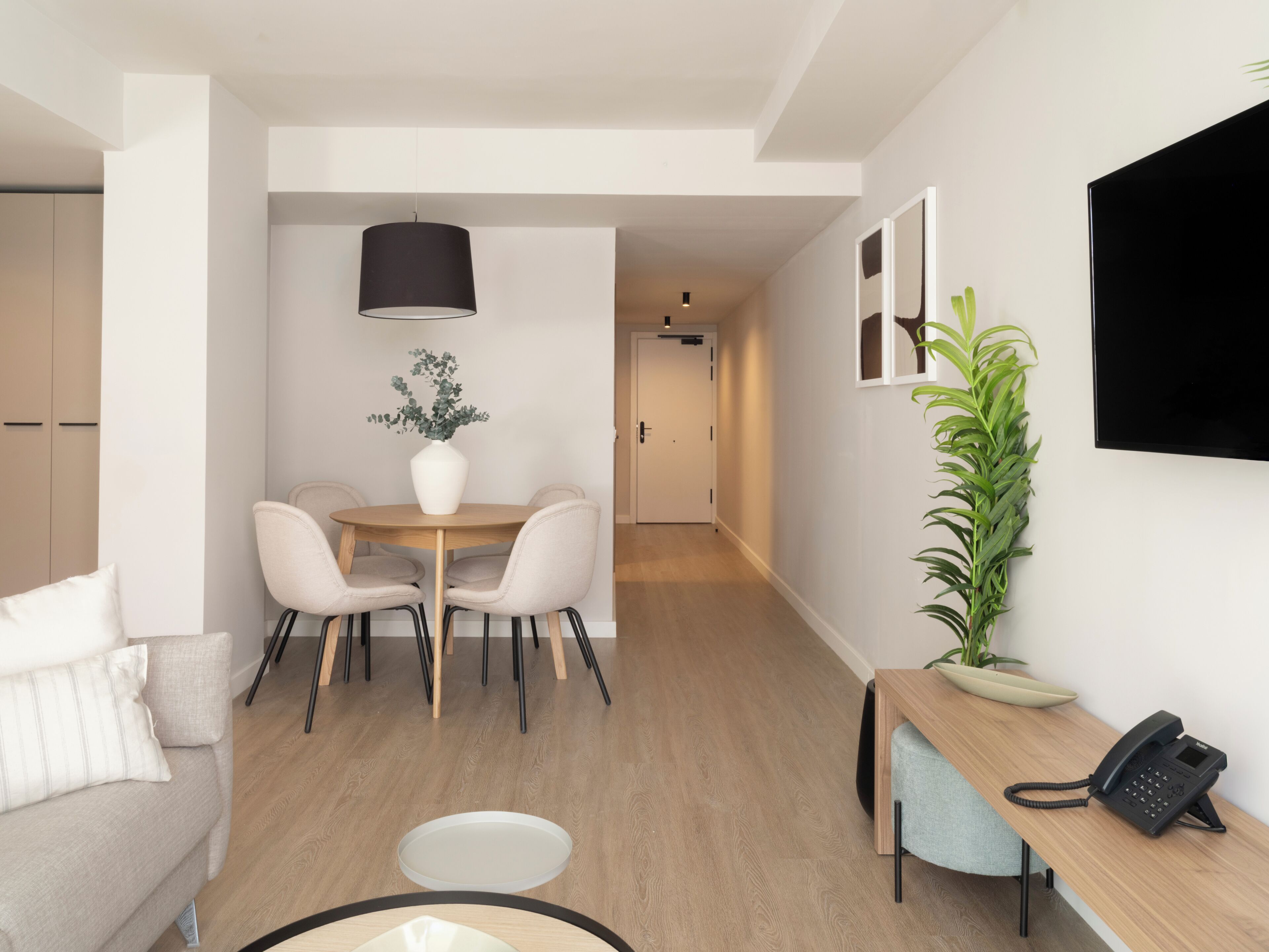 A modern hotel room with a dining area, a sofa, a desk with a phone, and a TV mounted on the wall.