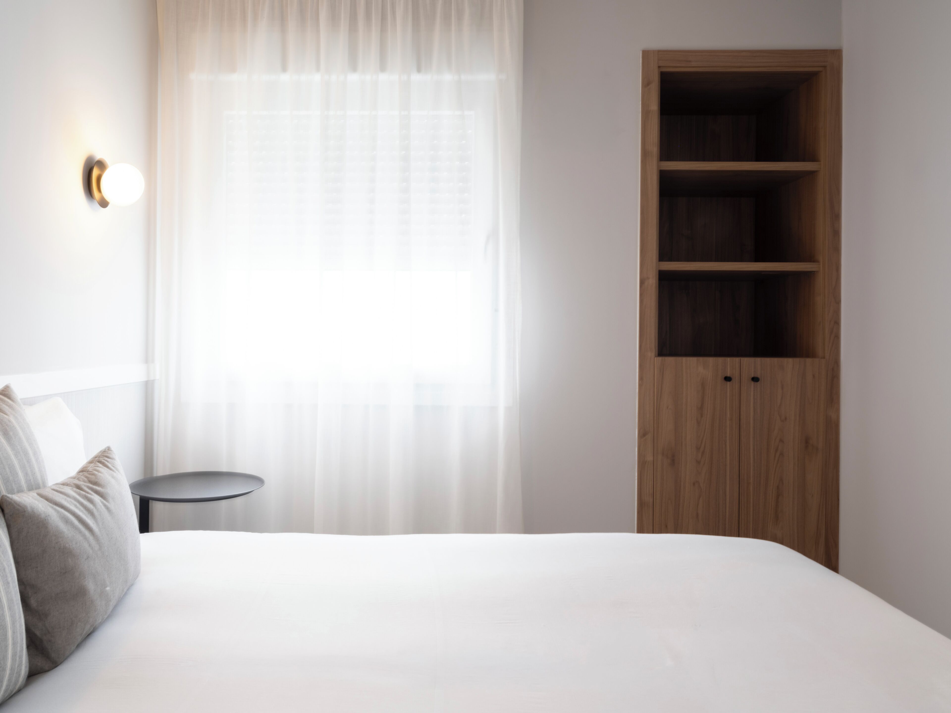 Un dormitorio con una cama, una mesita de noche, un armario de madera y una ventana con cortinas traslúcidas.