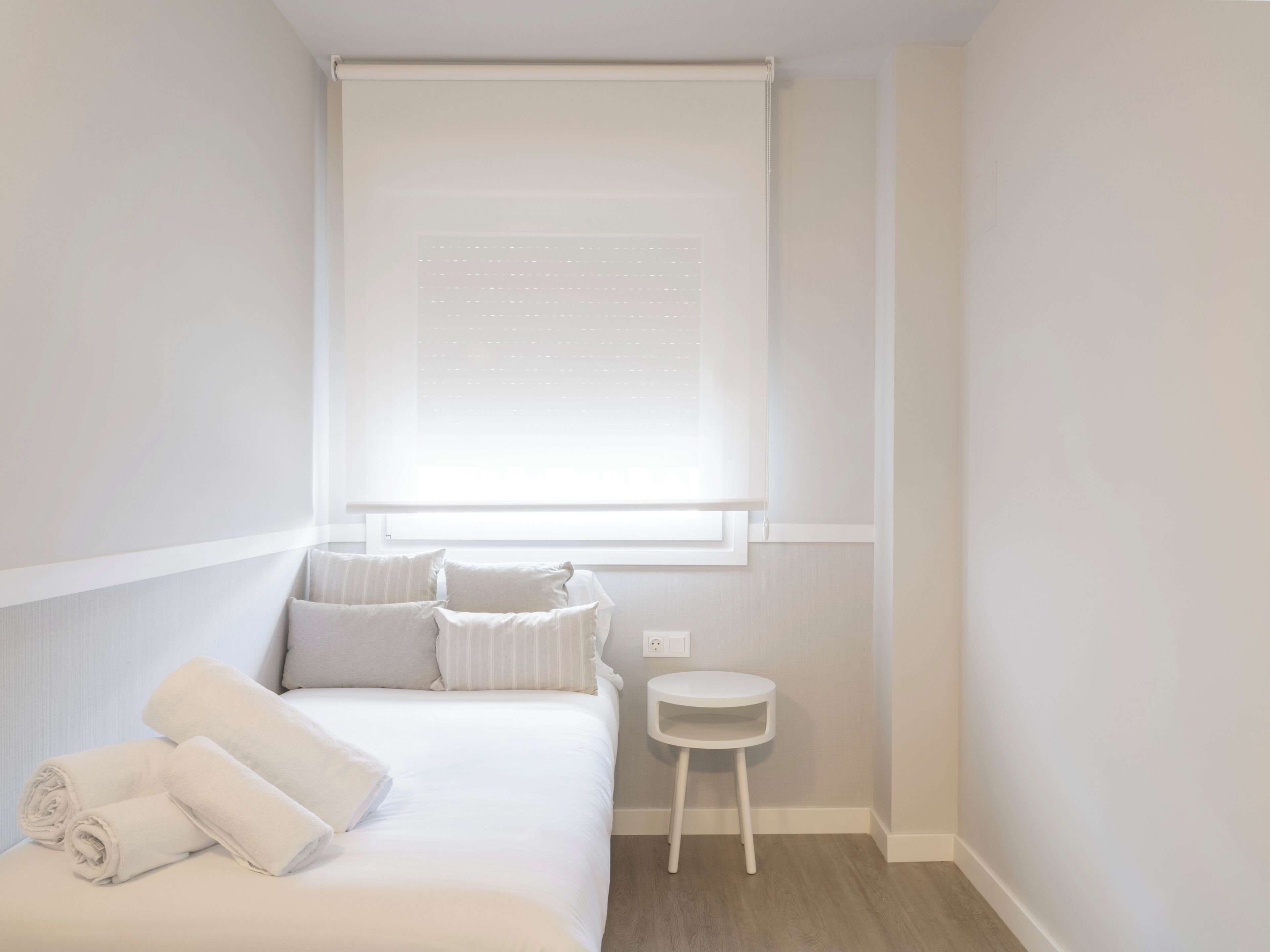 A bedroom with a bed, pillows, a small white stool, and a window with a blind.