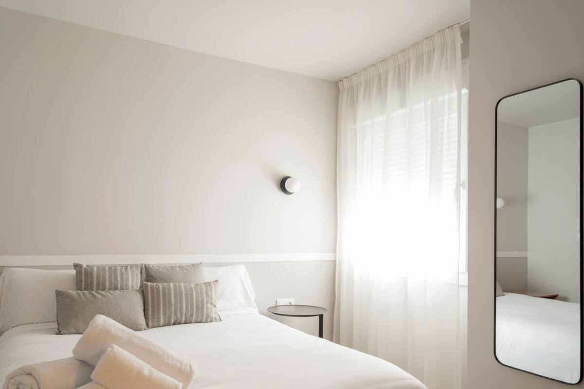 A hotel room with a neatly made bed, a round side table, a wall-mounted mirror, and sheer curtains.