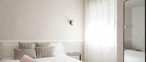 Une chambre d’hôtel avec un lit soigneusement fait, une petite table ronde, un miroir fixé au mur et des rideaux légers.