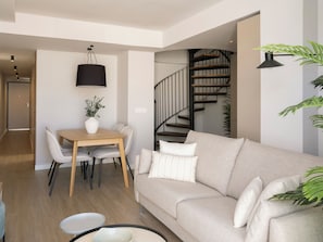 A modern living room with a sofa, a dining table with chairs, a staircase, and a hanging light fixture.