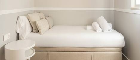 A small, single-bed room with a white bed, a small white table, and a window.