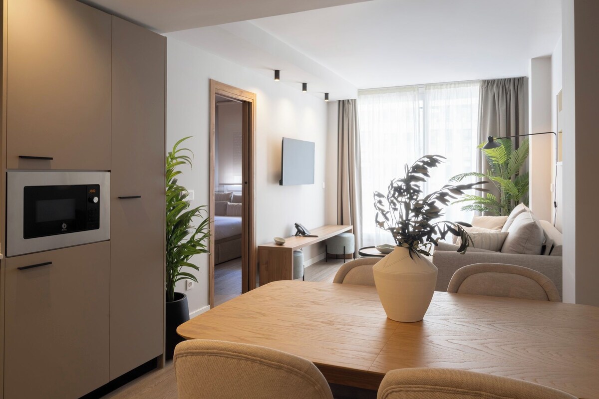 A modern living room with a dining table, a sofa, a TV mounted on the wall, and a potted plant.