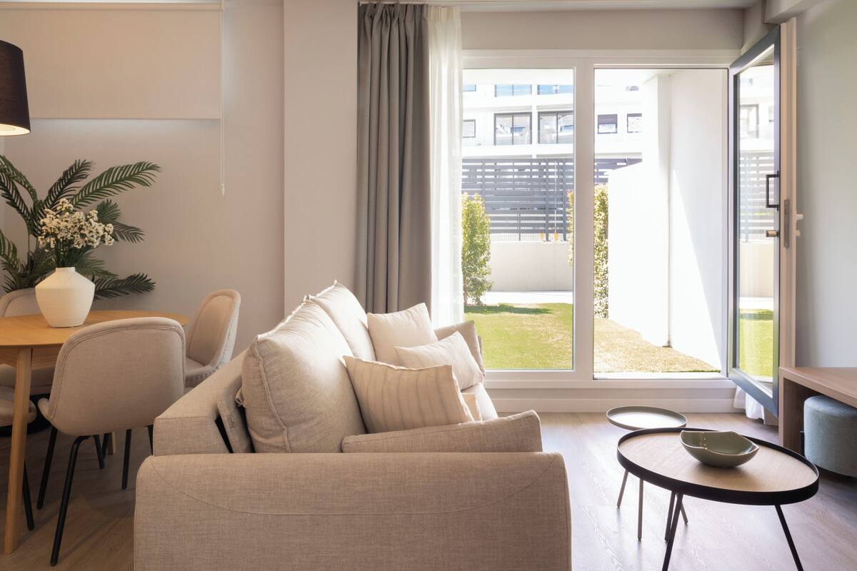 A modern living room with a sofa, a dining table, and a view of a building and greenery outside.