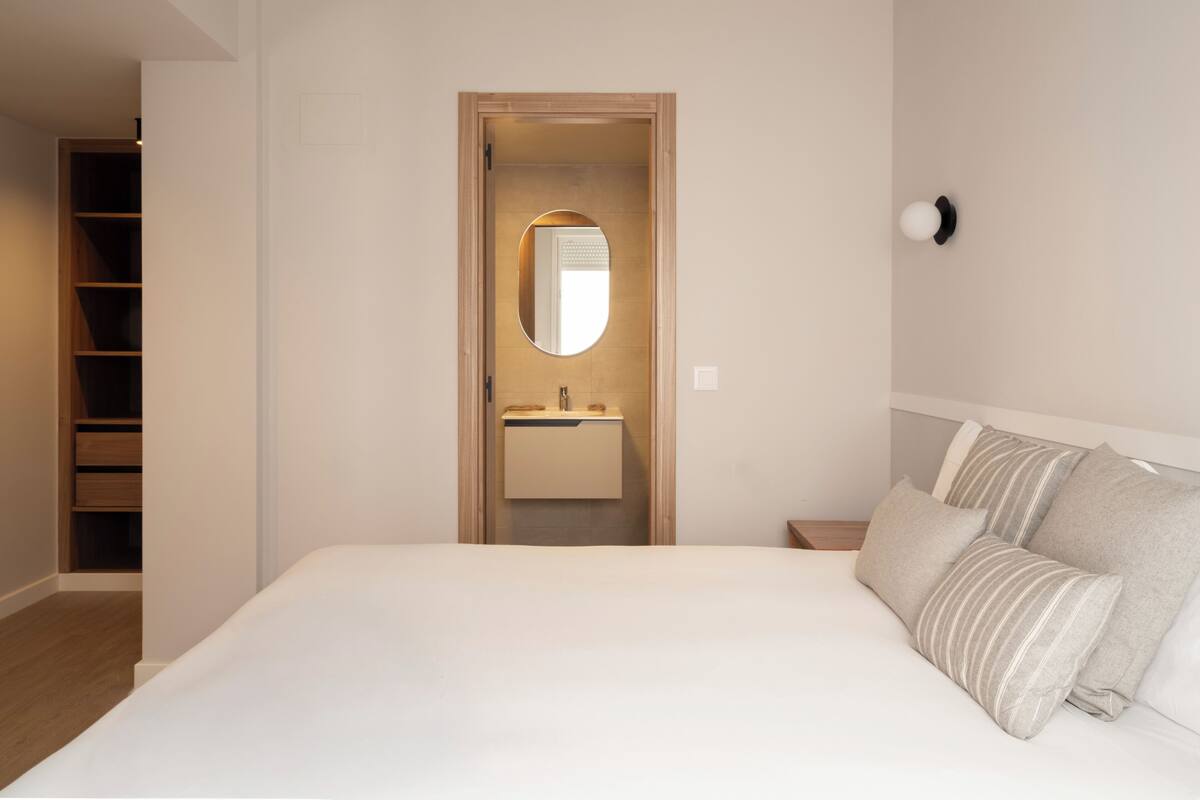 A bedroom with a bed, a bedside table, a nightstand, a wardrobe, and a bathroom with a mirror and sink.
