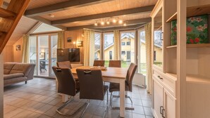 Un comedor con mesa y sillas de madera, televisor y ventanas amplias con cortinas.