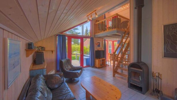 Interior acogedor de una cabaña con techo de madera, un sofá de cuero, una mesa de madera, chimenea, televisor y un balcón con escaleras.