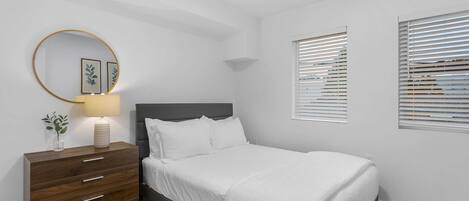 Ein Schlafzimmer mit einem Bett, einem Schminktisch, einem Spiegel und zwei Fenstern mit Jalousien.