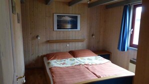 A wooden-paneled room with a bed, a small table, a wall-mounted shelf, and a window with curtains. - Ferienhaus im skandinavischen Stil (Granzow)