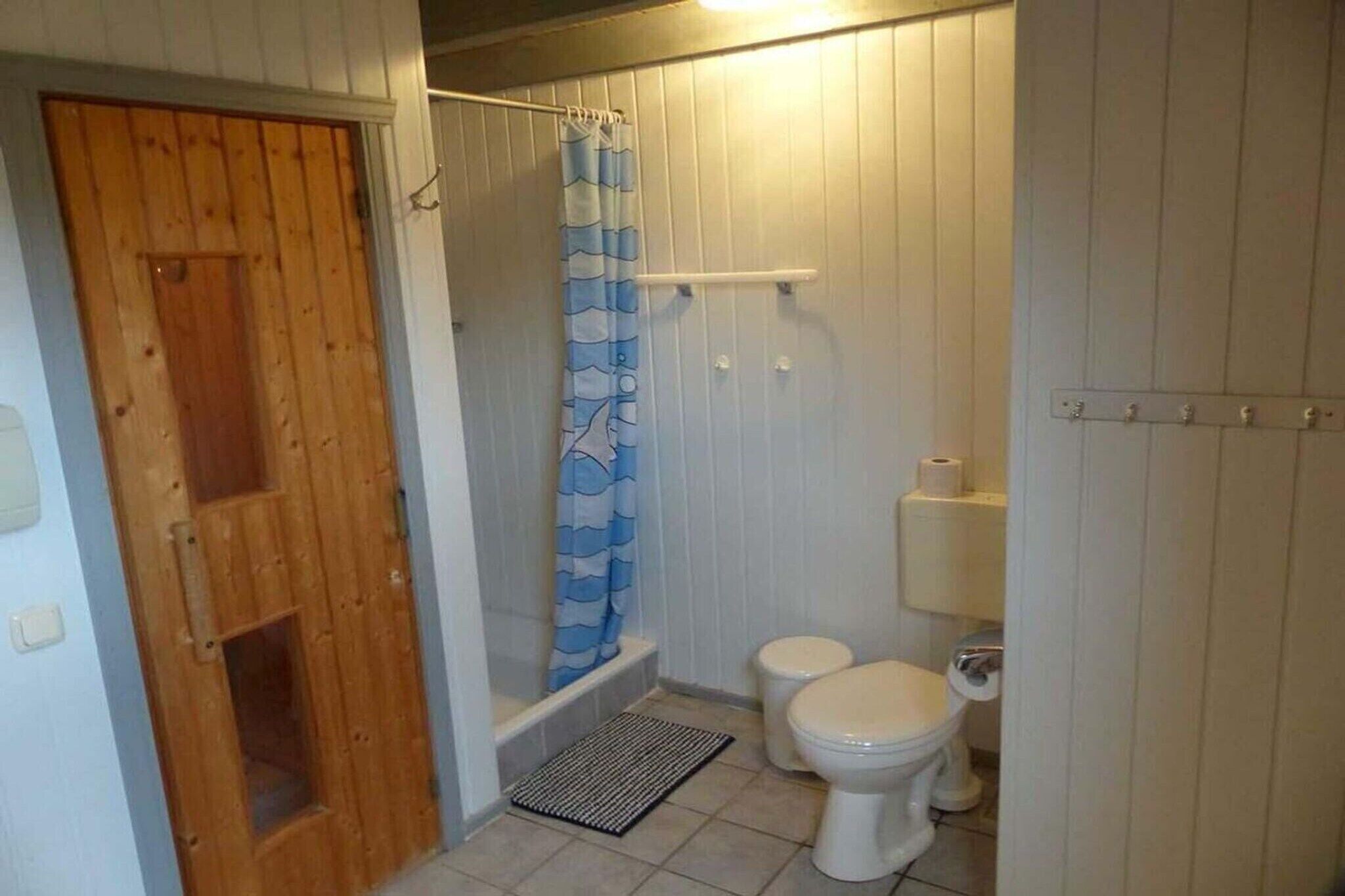 Un baño pequeño y compacto con puerta de madera, ducha con cortina azul y blanca, dos inodoros blancos y un lavamanos blanco.
