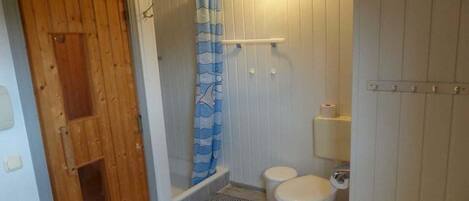Un baño pequeño y compacto con puerta de madera, ducha con cortina azul y blanca, dos inodoros blancos y un lavamanos blanco.