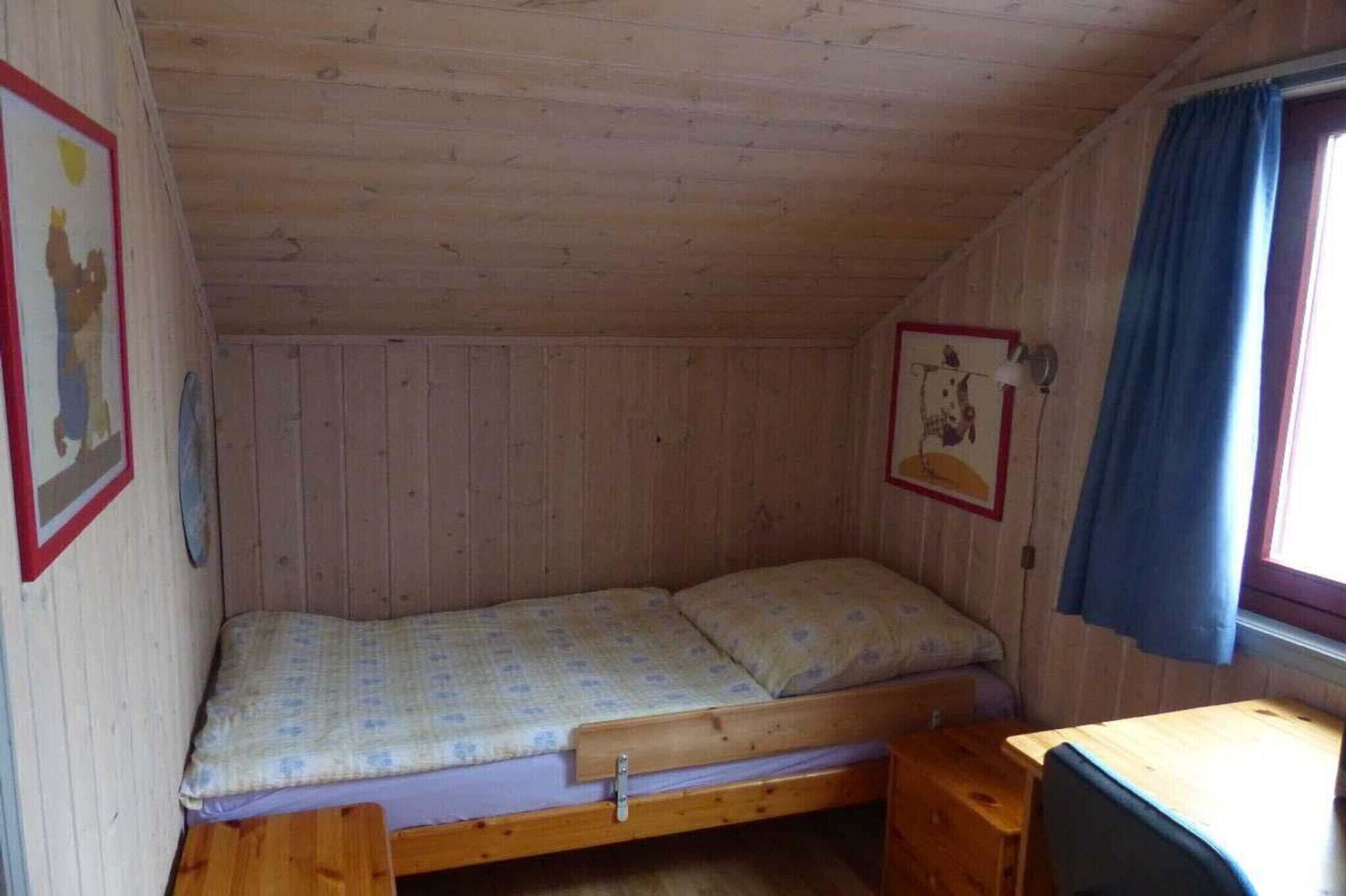 Ein kleines Zimmer mit einem Einzelbett, einem Holzbettkasten, einem Tisch und einem Fenster mit Vorhängen.