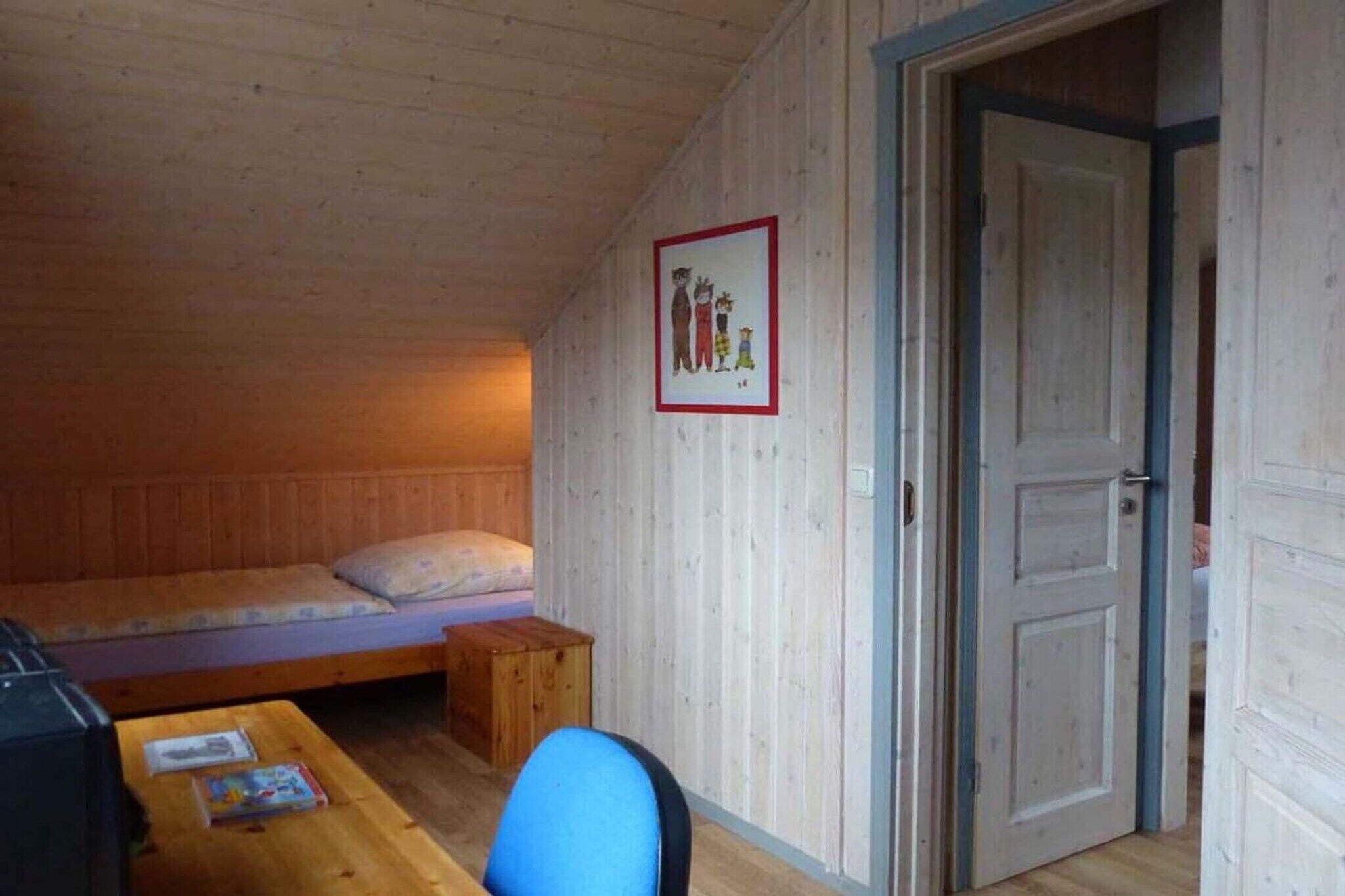A small, cozy room with a wooden ceiling and walls, a single bed, a small table with a chair, and a door leading to another room.