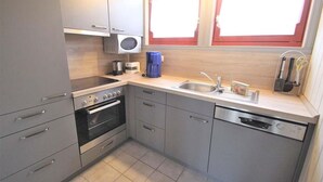 A compact kitchen with grey cabinets, a built-in oven, a dishwasher, and a window with a red frame. - Ferienhaus im skandinavischen Stil (Granzow)
