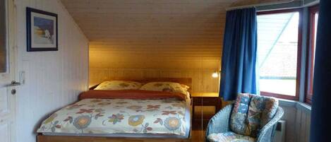Un dormitorio acogedor con techo de madera, una cama con ropa de cama floral, una silla de mimbre y una mesita redonda.