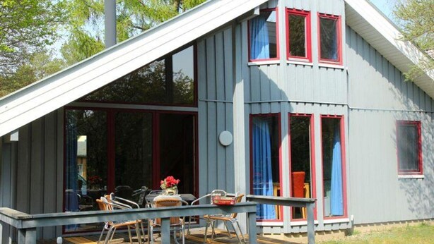 Een gebouw met twee verdiepingen, een schuin dak, grote ramen met rode omlijsting en een overdekt terras met tafels en stoelen.