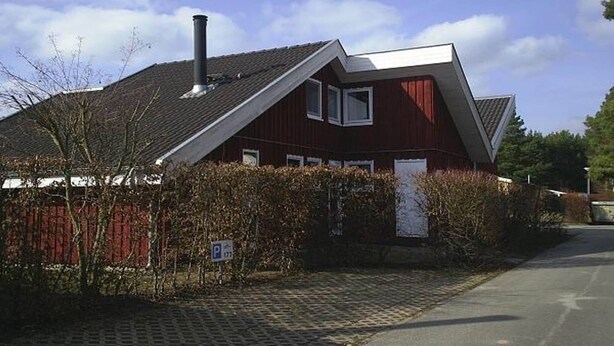 Una casa roja con techo inclinado, chimenea y un pequeño jardín con seto y entrada pavimentada.