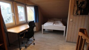 Una habitación con techo inclinado de madera, una cama, un escritorio con silla y una ventana con cortinas.