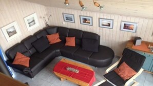 Sala de estar con un sofá seccional negro, un reposapiés rojo y una mesa de centro de madera. Se observan cuadros enmarcados en la pared y una mesita con una lámpara.