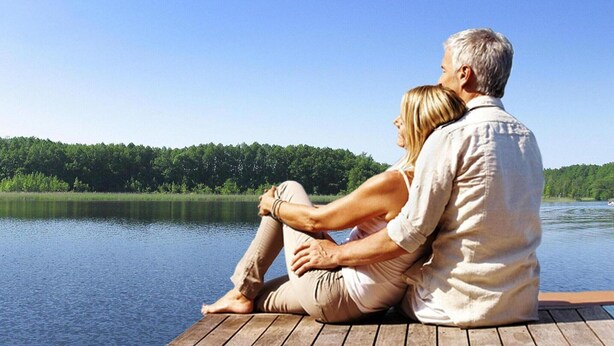 A couple sitting on a wooden dock overlooking a calm lake with a forested shoreline.
