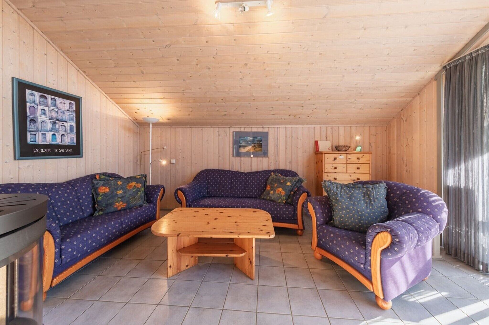 A cozy room with wooden walls and ceiling, featuring two purple sofas with floral cushions, a wooden coffee table, and a small wooden cabinet with drawers.
