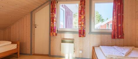 Una habitación con paredes de madera y techo inclinado, dos camas, una ventana con cortinas y un radiador.