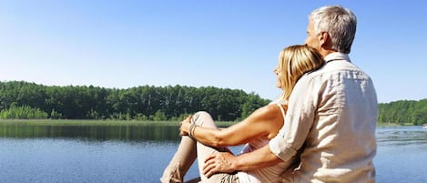 A couple sitting on a wooden dock overlooking a calm lake with a forested shoreline.