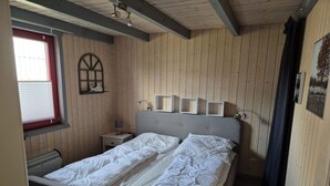 Un dormitorio con pared de madera, una cama con ropa de cama blanca, una mesita de noche con lámpara y una ventana con persianas.