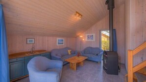 Un accogliente interno di una baita con soffitto in legno, due divani, un tavolo di legno, un angolo cottura e un camino a legna.