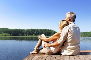 A couple sitting on a wooden dock overlooking a calm lake with a forested shoreline.