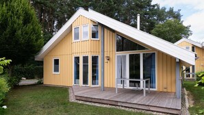Une maison en bois jaune, avec un toit en pente, de grandes fenêtres en verre et une terrasse en bois.